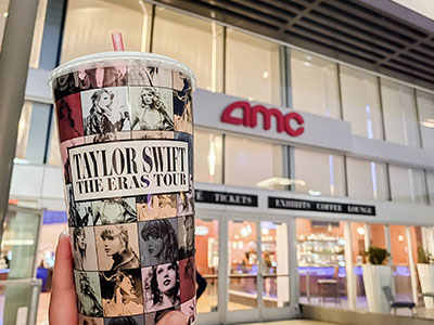 A person holding a beverage cup that says "Taylor Swift The Eras Tour" in front of a movie theater.