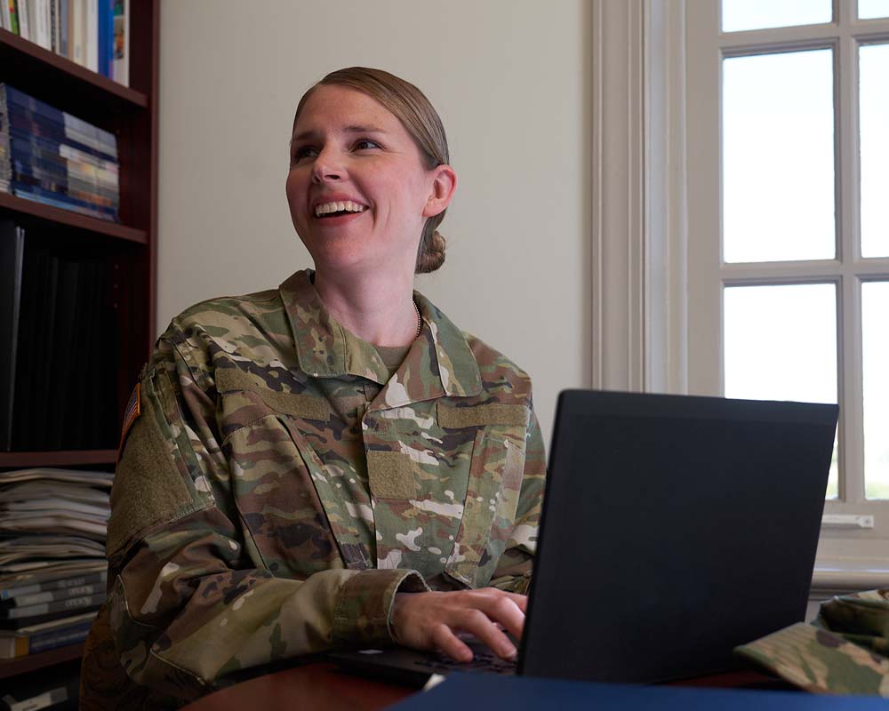 A person in a military uniform typing on a laptop and smiling.