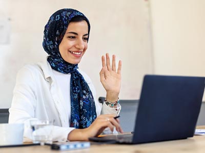Woman wearing a hijab working at a laptop and raising her hand.