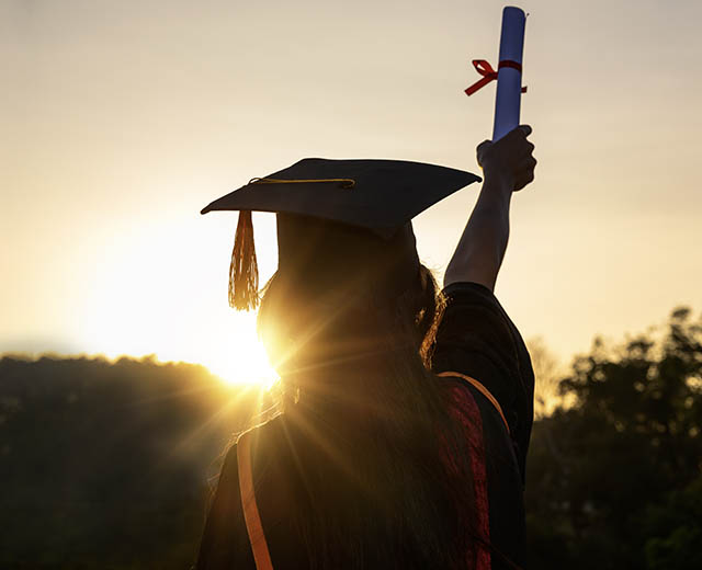 A person in graduation regalia holding a diploma in the air at sunset.