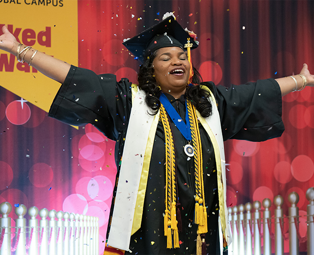 A UMGC graduate in graduation regalia with arms outstretched.