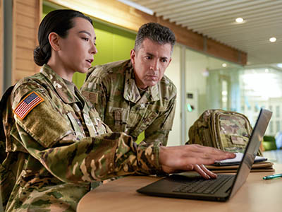 A woman in a military uniform sits at a laptop and points to the screen, while a man in military uniform stands next to her and looks at the laptop screen.