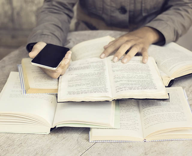 A person's hand on open books while holding a cell phone in the other hand.